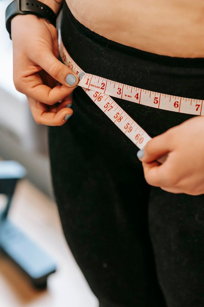 From above of crop anonymous plump female using measuring tape around hips in gym